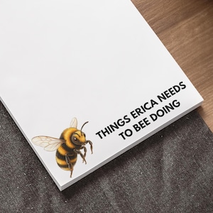 Może przedstawiać: Biały prostokątny notatnik z grafiką żółto-czarnego trzmiela w lewym dolnym rogu oraz czarnym napisem "THINGS ERICA NEEDS TO BEE DOING" wydrukowanym w prawym dolnym rogu na czystej, białej powierzchni papieru.