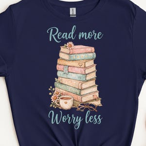 May include: Dark blue t-shirt with a print of a stack of books, a coffee cup, and glasses. The text "Read more Worry less" is in light blue script. The shirt has a crew neck.