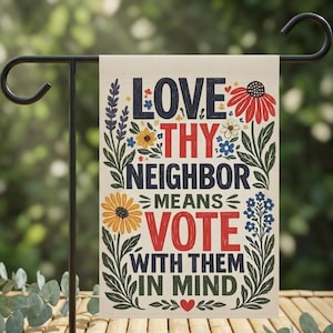 May include: A rectangular garden flag hanging on a black metal stand. The cream-colored fabric features the text LOVE THY NEIGHBOR MEANS VOTE WITH THEM IN MIND in dark blue and red, surrounded by illustrations of colorful flowers and green leaves.