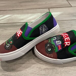May include: Pair of custom-painted slip-on shoes. The shoes feature a black and maroon toe with a green and purple trim. One shoe has a skull graphic and the word "VE" in red. The other shoe has a monster truck graphic and the word "GGER" in red.