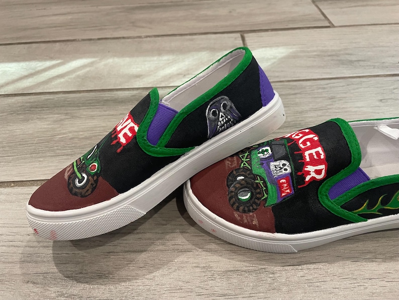 May include: Pair of custom-painted slip-on shoes. The shoes feature a black and maroon toe with a green and purple trim. One shoe has a skull graphic and the word "VE" in red. The other shoe has a monster truck graphic and the word "GGER" in red.
