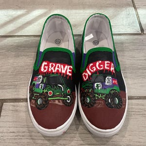 May include: Pair of slip-on shoes featuring a monster truck design. The shoes have a black upper with the words "GRAVE DIGGER" in red and purple. The monster truck is green and black with a skull graphic. The shoes have a white sole and a green trim.
