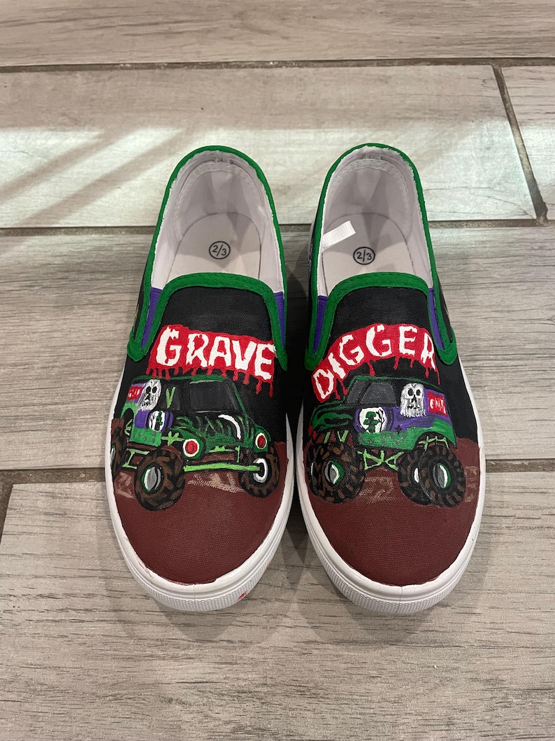 May include: Pair of slip-on shoes featuring a monster truck design. The shoes have a black upper with the words "GRAVE DIGGER" in red and purple. The monster truck is green and black with a skull graphic. The shoes have a white sole and a green trim.