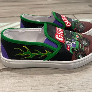 May include: A pair of slip-on shoes with a monster truck theme. The shoes feature a black and purple design with green flames and the words "GRAVE DIGGER" and "DIGGER" in red. The shoes have white soles.