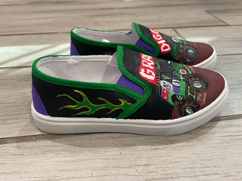 May include: A pair of slip-on shoes with a monster truck theme. The shoes feature a black and purple design with green flames and the words "GRAVE DIGGER" and "DIGGER" in red. The shoes have white soles.