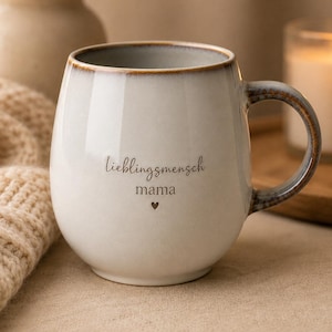 Könnte beinhalten: Eine bauchige Keramiktasse mit weißer Glasur und dunkelbraunem Rand. Der Schriftzug "lieblingsmensch" und "mama" ist in Schwarz aufgedruckt, gefolgt von einem kleinen Herzsymbol. Der Henkel ist geschwungen und farblich auf den Rand abgestimmt.