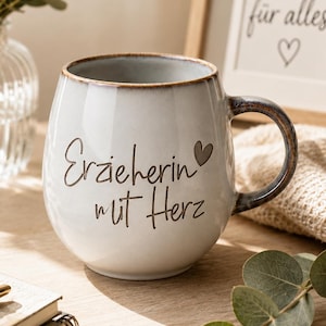 May include: A round ceramic mug with a light gray glaze and a dark brown rim. The mug features the dark brown text Erzieherin mit Herz and a small heart icon. The handle is dark brown with a mottled finish, and the base is rounded and smooth.