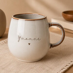 May include: A rounded ceramic mug with a cream-colored glaze and a brown speckled rim. The mug features the name Yvonne written in a thin, dark script font, followed by a small heart symbol. It has a curved handle with a brown, textured finish.