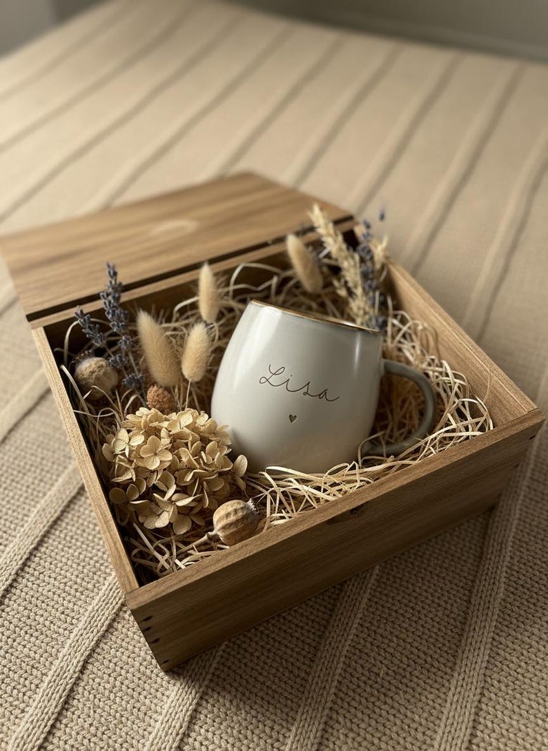 May include: A wooden gift box with a light gray mug that has the name "Lisa" written on it in cursive with a small heart. The mug has a gold rim and is surrounded by dried flowers and straw. The box is open.