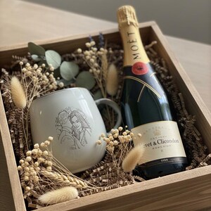 May include: A wooden gift box containing a bottle of Mo&euml;t & Chandon champagne and a light-colored ceramic mug featuring a line-art illustration of a couple. The box is filled with brown paper filler, dried eucalyptus, and fluffy beige grasses.