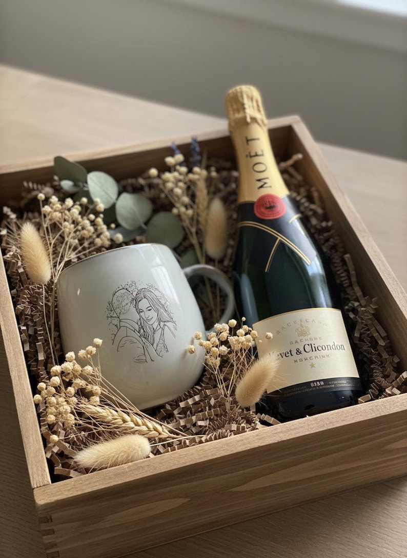 May include: A wooden gift box containing a bottle of Mo&euml;t & Chandon champagne and a light-colored ceramic mug featuring a line-art illustration of a couple. The box is filled with brown paper filler, dried eucalyptus, and fluffy beige grasses.