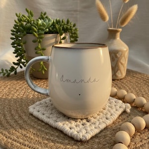 May include: Cream-colored ceramic mug with the name "Amanda" in script and a small heart. The mug sits on a square, off-white crocheted coaster, with a jute placemat underneath. A small vase with dried flowers and a potted succulent are in the background.