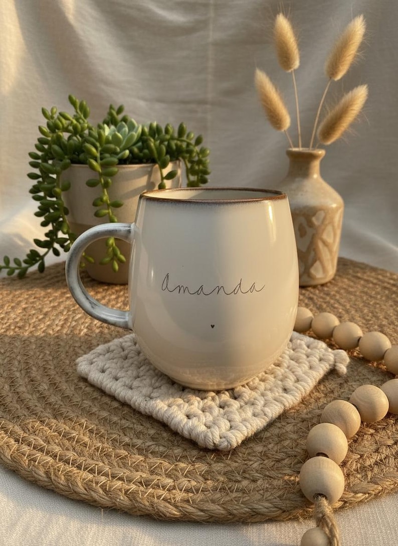 May include: Cream-colored ceramic mug with the name "Amanda" in script and a small heart. The mug sits on a square, off-white crocheted coaster, with a jute placemat underneath. A small vase with dried flowers and a potted succulent are in the background.