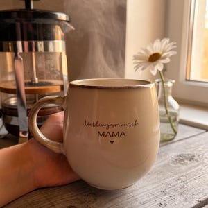 May include: A beige ceramic mug with the words "lieblingsmensch MAMA" and a heart symbol. Steam rises from the mug, suggesting a hot drink. A French press and a vase with a daisy are in the background.