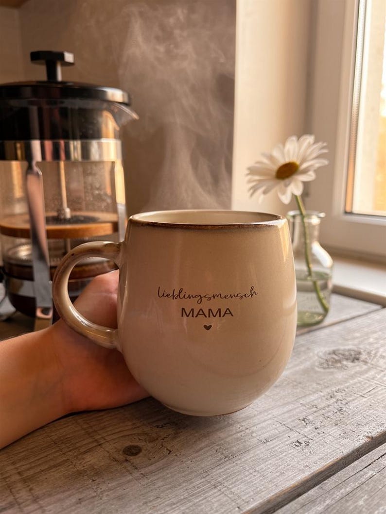 May include: A beige ceramic mug with the words "lieblingsmensch MAMA" and a heart symbol. Steam rises from the mug, suggesting a hot drink. A French press and a vase with a daisy are in the background.