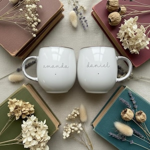 May include: Two white ceramic mugs with the names "amanda" and "daniel" in black script. The mugs are surrounded by vintage books in shades of burgundy, green, and teal, adorned with dried flowers and grasses. A cozy, rustic aesthetic.