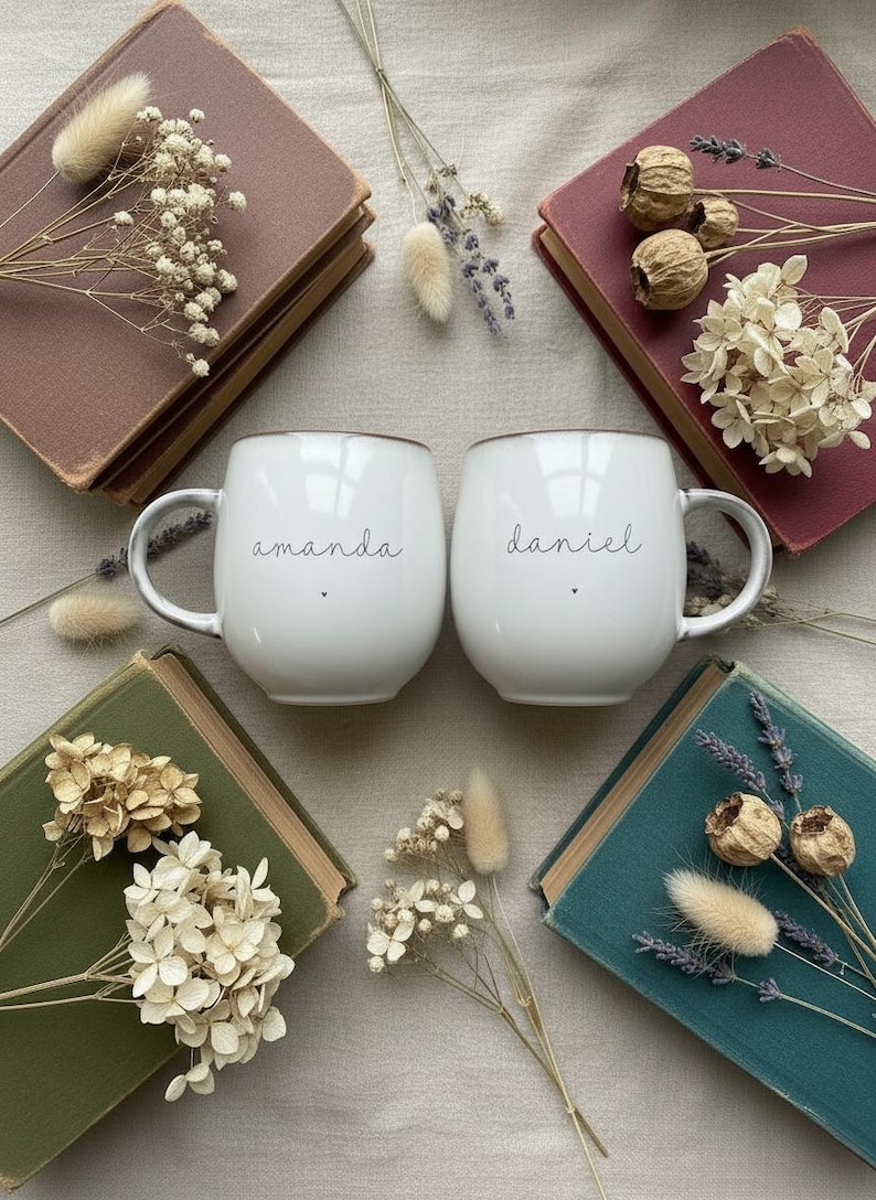 May include: Two white ceramic mugs with the names "amanda" and "daniel" in black script. The mugs are surrounded by vintage books in shades of burgundy, green, and teal, adorned with dried flowers and grasses. A cozy, rustic aesthetic.