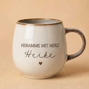 May include: A ceramic mug with a rounded shape, featuring a cream-colored glaze with a brown rim and handle. The text HEBAMME MIT HERZ and the name Heike are printed in black, accompanied by a small brown heart icon centered below the name.