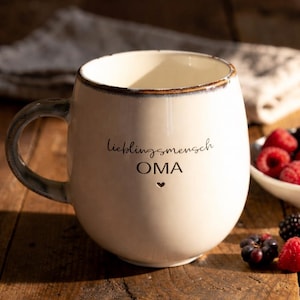 May include: Cream ceramic mug with a rounded shape and a dark handle. The mug has the words "lieblingsmensch OMA" and a heart symbol. Berries are visible in a small white bowl.