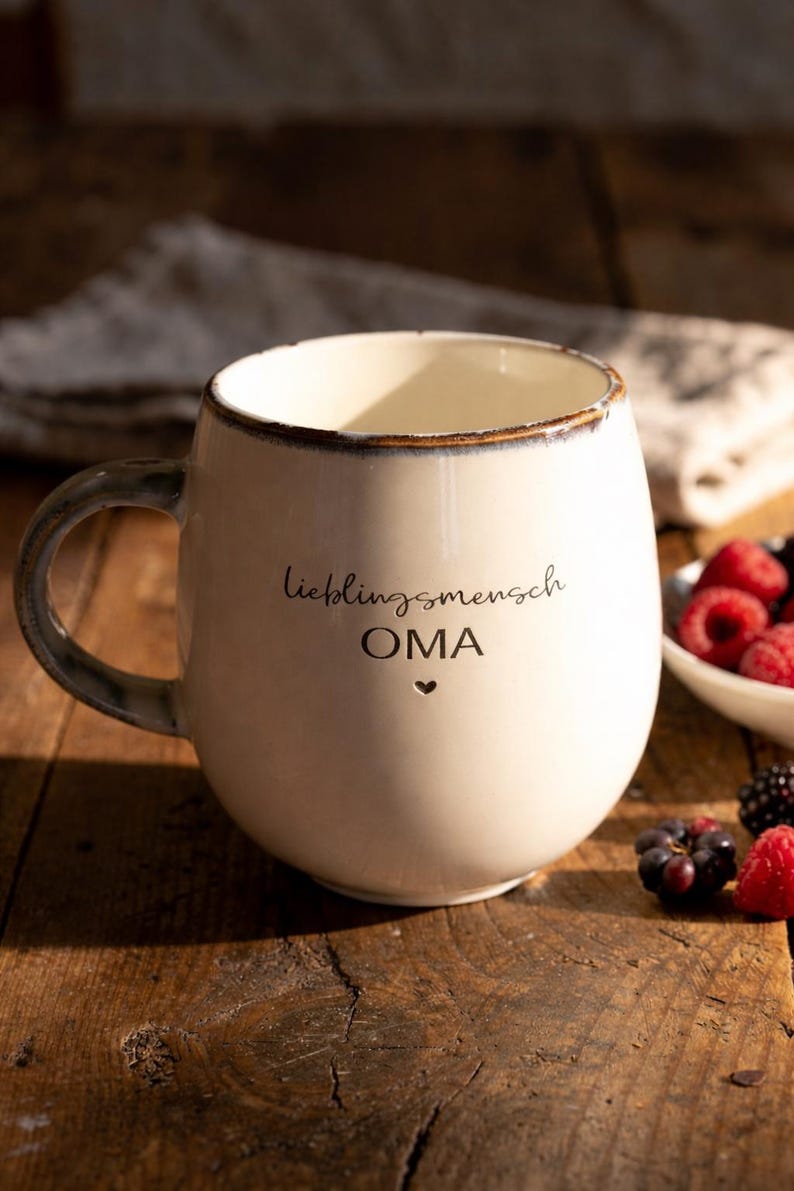 May include: Cream ceramic mug with a rounded shape and a dark handle. The mug has the words "lieblingsmensch OMA" and a heart symbol. Berries are visible in a small white bowl.