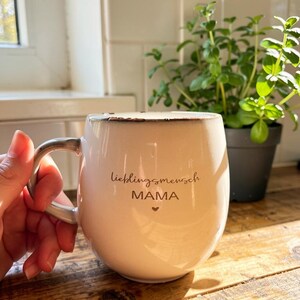 May include: Cream ceramic mug with the words "lieblingsmensch MAMA" and a heart. The mug has a silver handle and sits on a wooden surface. A small potted plant is in the background.