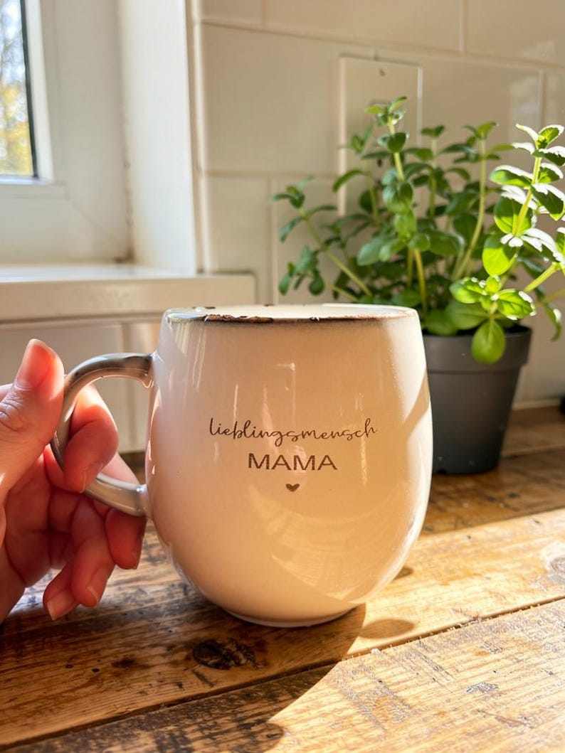 May include: Cream ceramic mug with the words "lieblingsmensch MAMA" and a heart. The mug has a silver handle and sits on a wooden surface. A small potted plant is in the background.