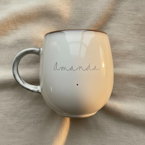 May include: White ceramic mug with a rounded shape and a gray handle. The word "Amanda" is written in cursive on the front, with a small heart below. The rim has a brown accent.