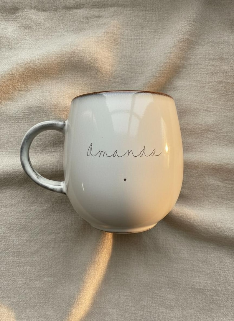 May include: White ceramic mug with a rounded shape and a gray handle. The word "Amanda" is written in cursive on the front, with a small heart below. The rim has a brown accent.