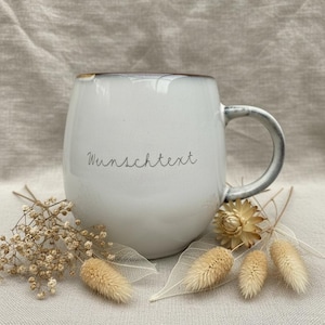 May include: White ceramic mug with a rounded shape and a gray handle. The mug has the word "Wunschtext" written on it. The mug is surrounded by dried flowers and decorative elements, creating a rustic aesthetic.