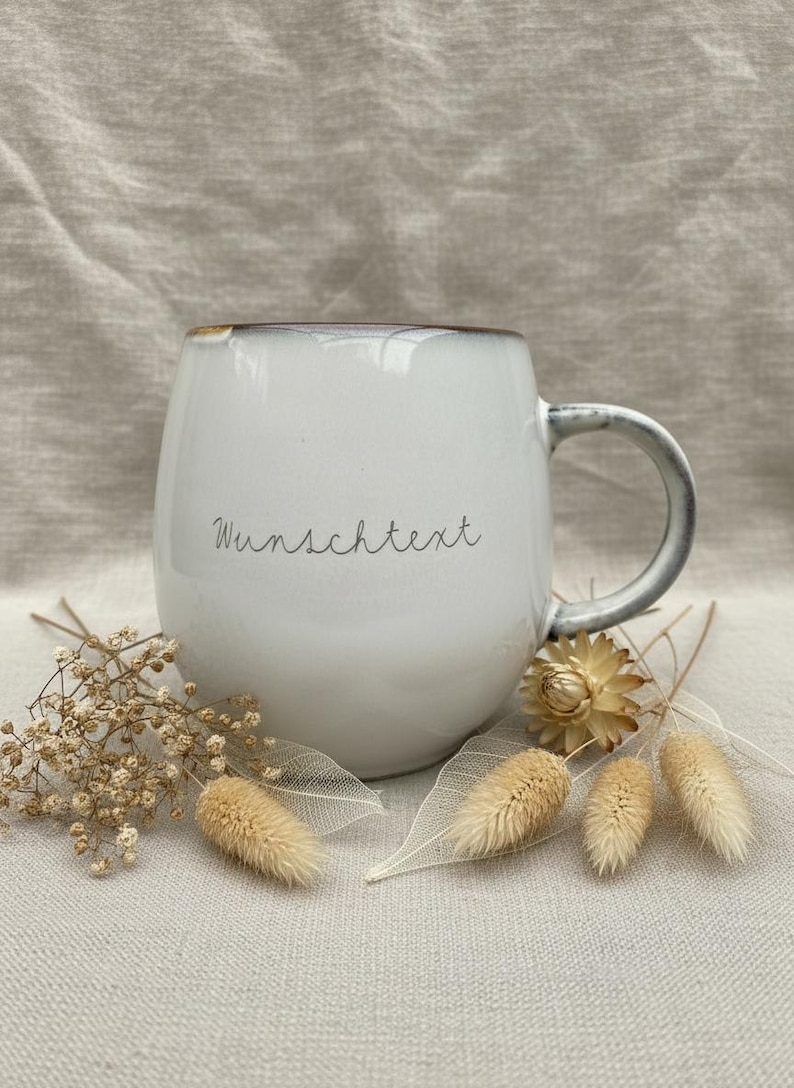 May include: White ceramic mug with a rounded shape and a gray handle. The mug has the word "Wunschtext" written on it. The mug is surrounded by dried flowers and decorative elements, creating a rustic aesthetic.