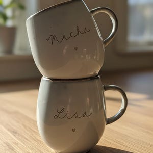 May include: Two stacked, off-white ceramic mugs with brown rims and handles. The top mug has the name "Micha" and a heart, while the bottom mug has "Lisa" and a heart. The mugs are on a wooden surface.