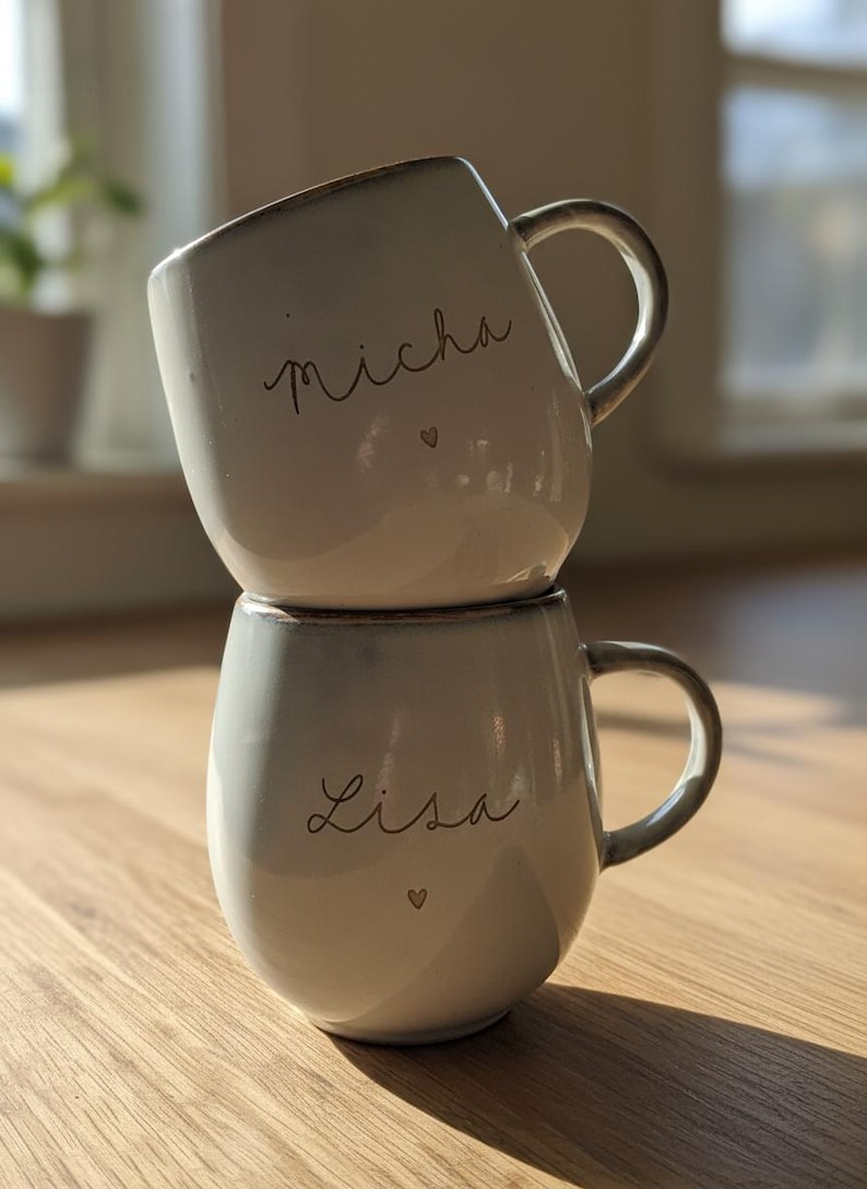 May include: Two stacked, off-white ceramic mugs with brown rims and handles. The top mug has the name "Micha" and a heart, while the bottom mug has "Lisa" and a heart. The mugs are on a wooden surface.
