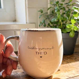May include: Cream ceramic mug with a distressed rim and a curved handle. The mug has the German text "Lieblingsmensch" and the name "THEO" with a heart symbol. A hand holds the mug, with a plant in the background.