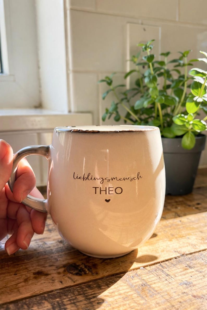 May include: Cream ceramic mug with a distressed rim and a curved handle. The mug has the German text "Lieblingsmensch" and the name "THEO" with a heart symbol. A hand holds the mug, with a plant in the background.