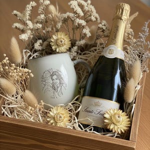 May include: A wooden gift box containing a white ceramic mug with a line-art illustration of a couple, a bottle of sparkling wine with a gold foil neck, and dried cream-colored flowers and grasses nestled in shredded paper filler.