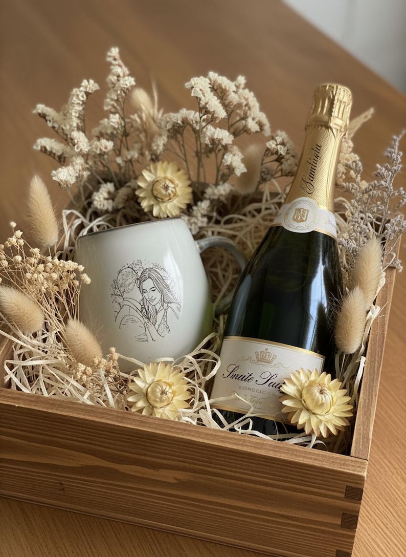 May include: A wooden gift box containing a white ceramic mug with a line-art illustration of a couple, a bottle of sparkling wine with a gold foil neck, and dried cream-colored flowers and grasses nestled in shredded paper filler.