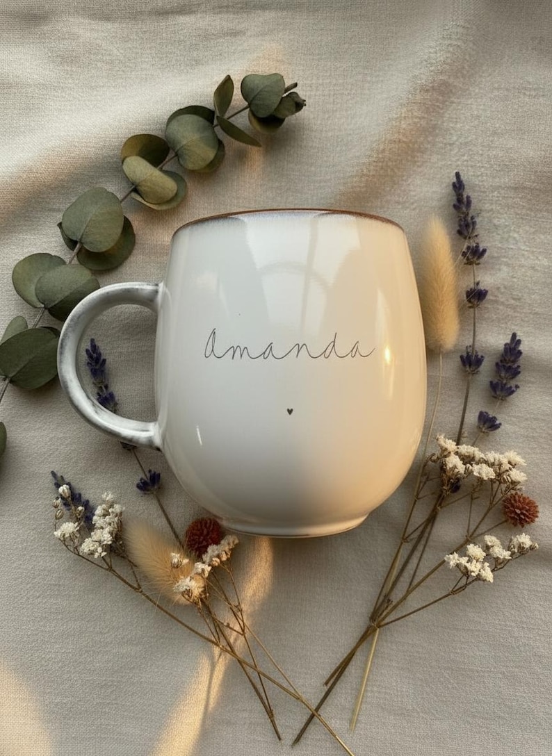 May include: White ceramic mug with the name "amanda" in a handwritten font and a small heart. The mug has a brown rim and a curved handle. The mug is surrounded by dried flowers and greenery.