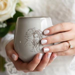 May include: A light gray ceramic mug featuring a dark brown line art illustration of a couple embracing. The mug has a smooth, rounded shape with a subtle gradient near the rim, showcasing a detailed sketch of two people in a close, intimate pose.
