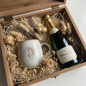May include: A wooden gift box containing a white ceramic mug with a line-art illustration of a couple, a bottle of Chateau Dore Brut Reserve sparkling wine, and dried floral accents including roses and bunny tails nestled in wood shavings.