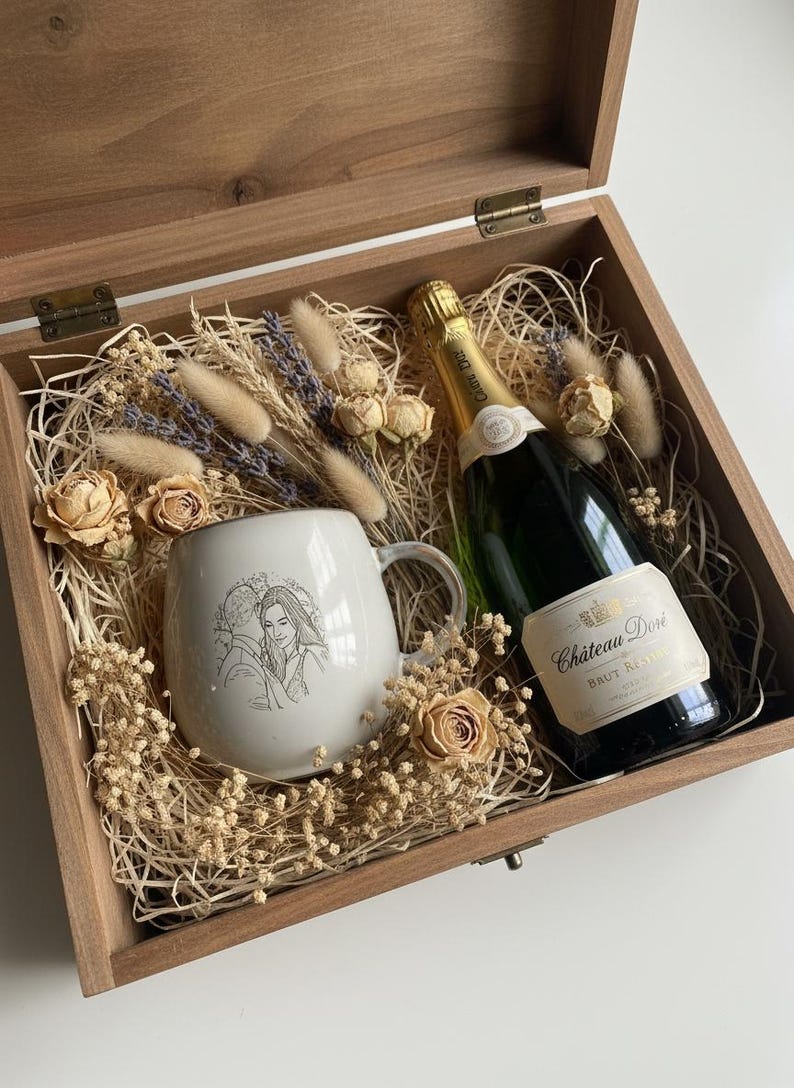 May include: A wooden gift box containing a white ceramic mug with a line-art illustration of a couple, a bottle of Chateau Dore Brut Reserve sparkling wine, and dried floral accents including roses and bunny tails nestled in wood shavings.