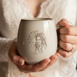 May include: A light gray ceramic mug with a rounded shape and a smooth finish. The front of the mug features a detailed brown line art illustration of a couple embracing. The mug has a sturdy handle on the right side and a subtle rim detail.