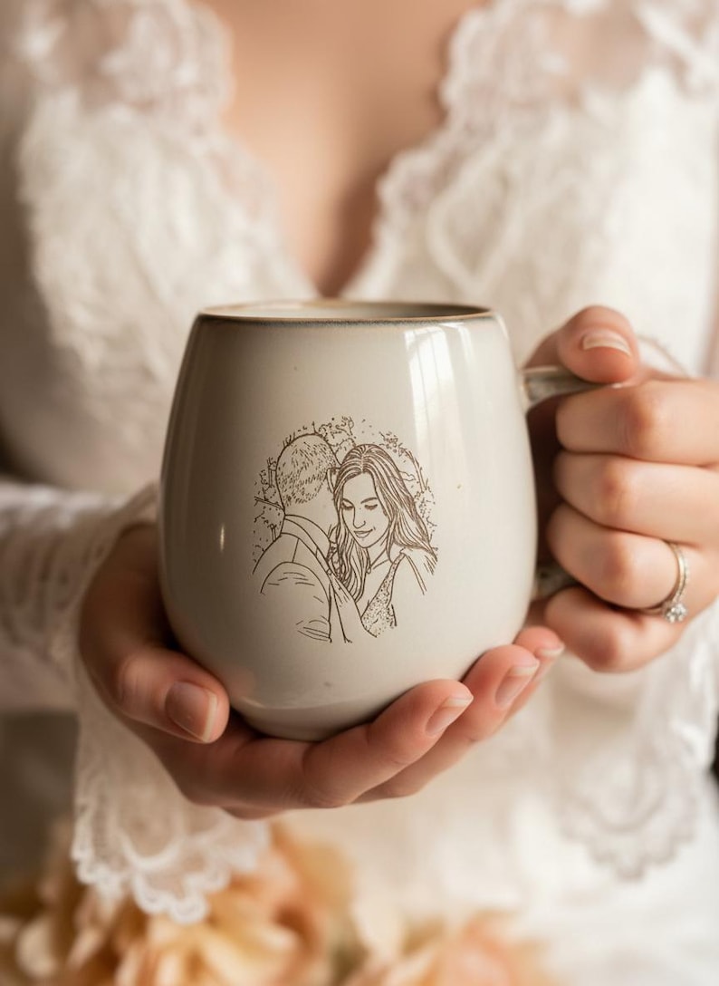 May include: A light gray ceramic mug with a rounded shape and a smooth finish. The front of the mug features a detailed brown line art illustration of a couple embracing. The mug has a sturdy handle on the right side and a subtle rim detail.