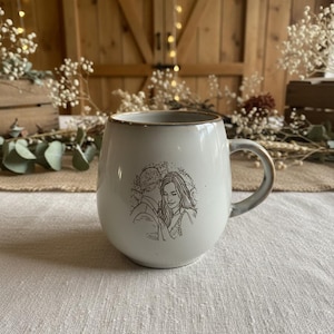 May include: A round, off-white ceramic mug featuring a dark brown line art illustration of two people standing close together. The mug has a smooth, glossy finish with a slightly darker rim and a curved handle on the right side of the vessel.