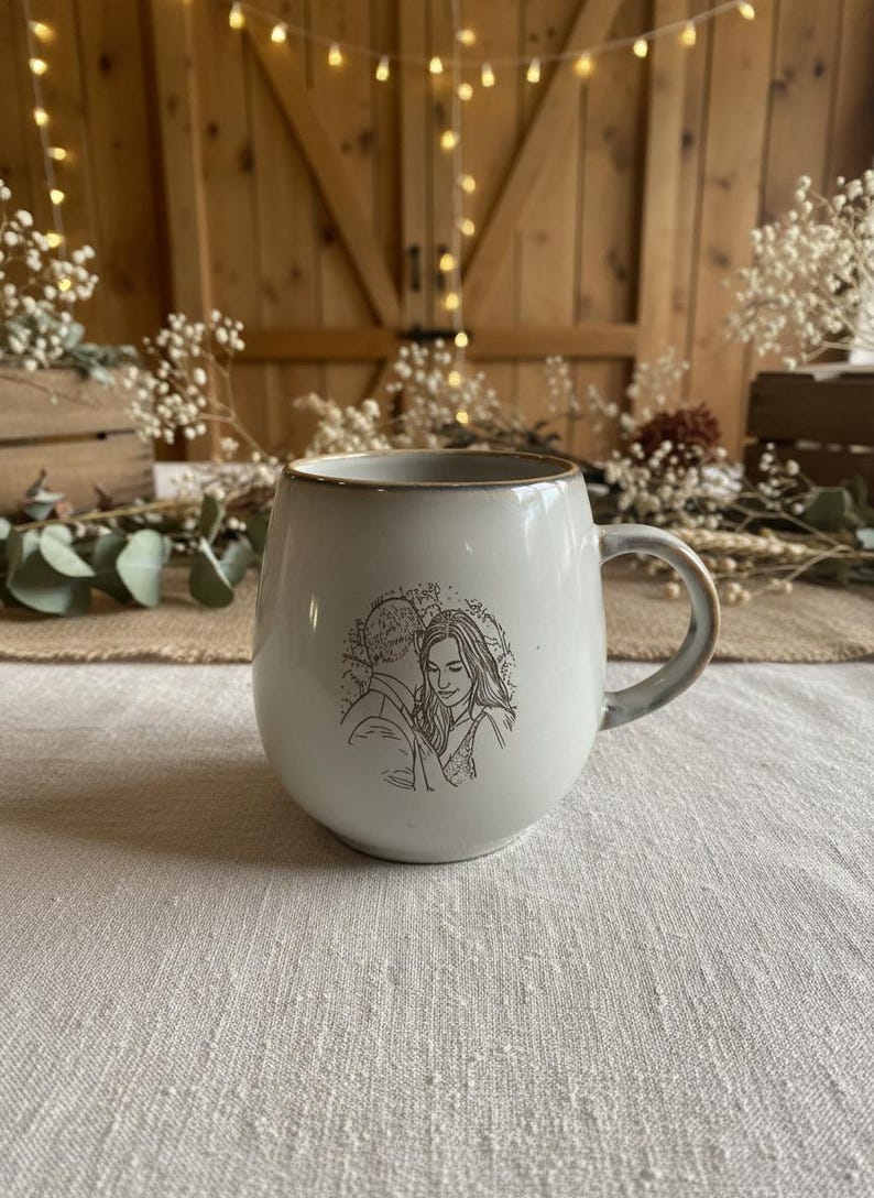 May include: A round, off-white ceramic mug featuring a dark brown line art illustration of two people standing close together. The mug has a smooth, glossy finish with a slightly darker rim and a curved handle on the right side of the vessel.
