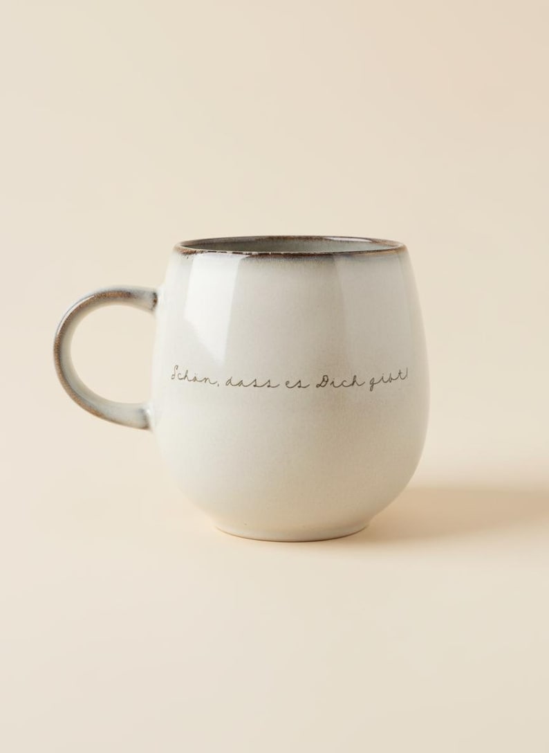 May include: A white ceramic mug with a rounded shape and a dark brown rim. The mug has a curved handle and features the handwritten text "Sch&ouml;n, dass es Dich gibt!" in black. A simple and elegant design.