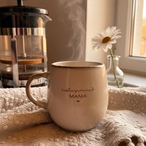 May include: A beige ceramic mug with the words "lieblingsmensch MAMA" printed on it, with steam rising from the contents. A French press and a daisy in a vase are in the background. The mug sits on a textured, fringed cloth.