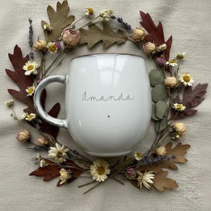 May include: White ceramic mug with the name "Amanda" in a delicate script, accented by a small heart. The mug is surrounded by a wreath of dried flowers and leaves, creating a rustic, natural aesthetic.