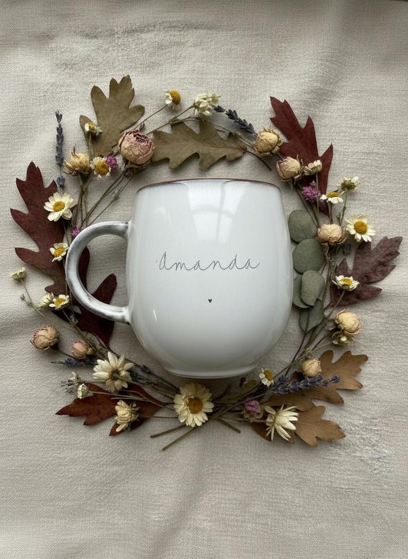 May include: White ceramic mug with the name "Amanda" in a delicate script, accented by a small heart. The mug is surrounded by a wreath of dried flowers and leaves, creating a rustic, natural aesthetic.