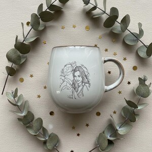 May include: A white ceramic mug with a rounded body and a handle, featuring a black line art illustration of two people embracing on the front. The mug is surrounded by two curved eucalyptus branches and small gold star and circle confetti.