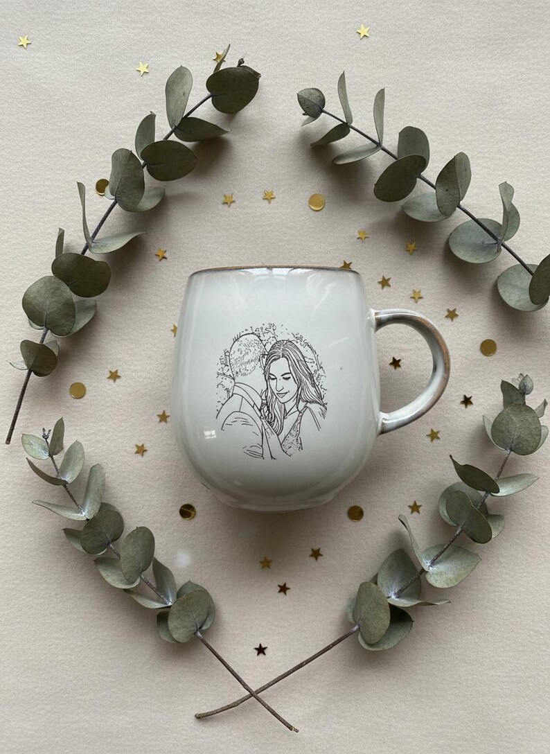 May include: A white ceramic mug with a rounded body and a handle, featuring a black line art illustration of two people embracing on the front. The mug is surrounded by two curved eucalyptus branches and small gold star and circle confetti.